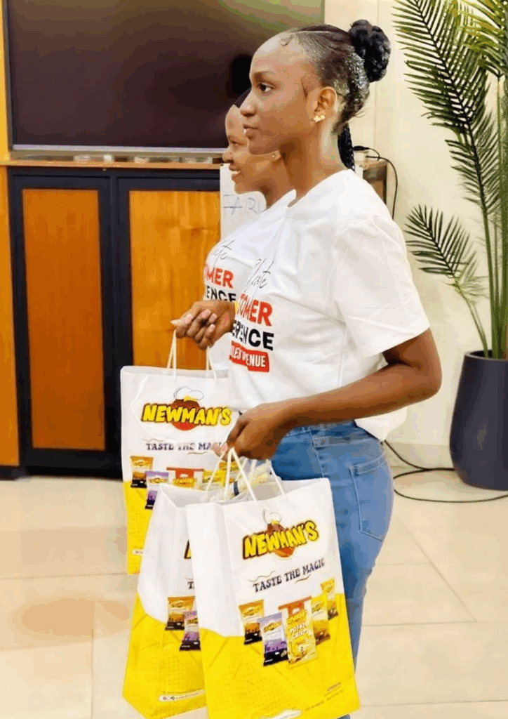 Two African women in white branded t-shirts and jeans standing, holding large white and yellow Newman's gift bags that read "Taste the Magic."