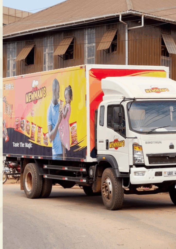 A white Sinotruk delivery truck, branded with a large Newman's advertisement on the side. The ad features a man in a blue shirt sharing a bag of Newman's chips with a little girl in a pink dress, and the slogan "Taste The Magic." The truck is parked on a street in front of a colonial-style building.
