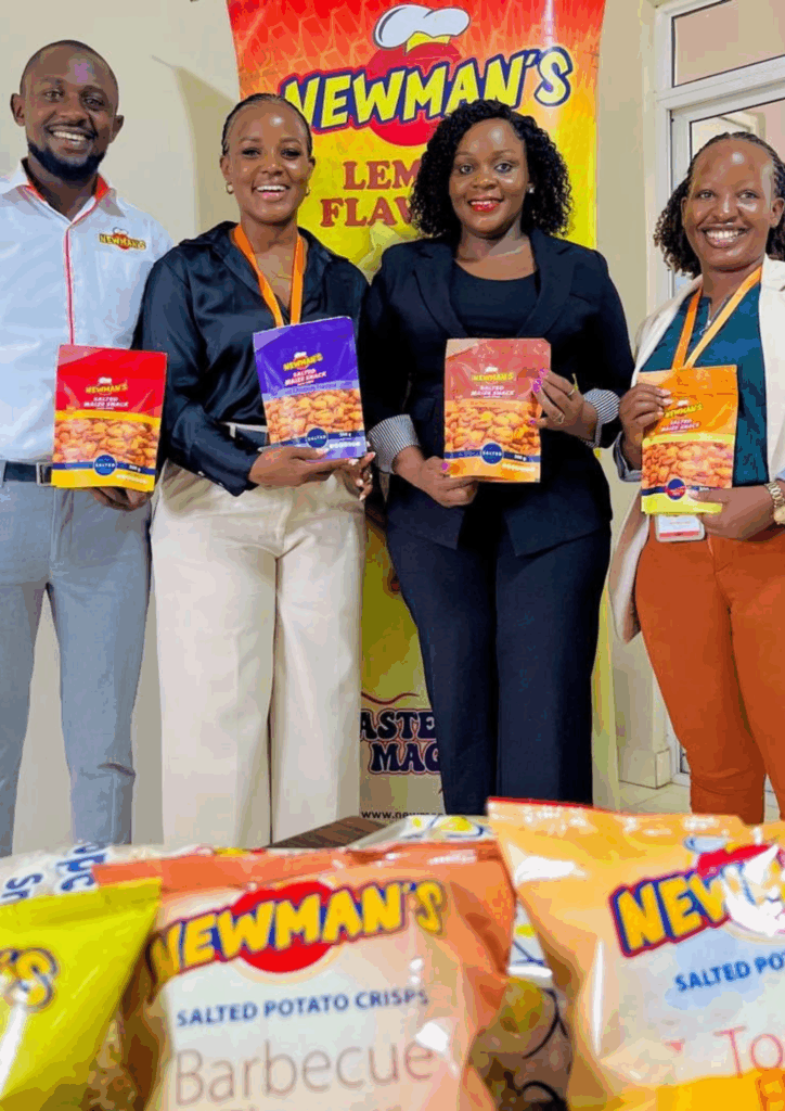 A group of four smiling people (three women and one man) stand behind a table laden with bags of chips. The people in the photo are holding up bags of Newman's chips in different flavors, including yellow (possibly Lemon flavor, which is visible on the banner), red, and orange packaging. A large yellow and red banner with the Newman's logo is displayed behind them