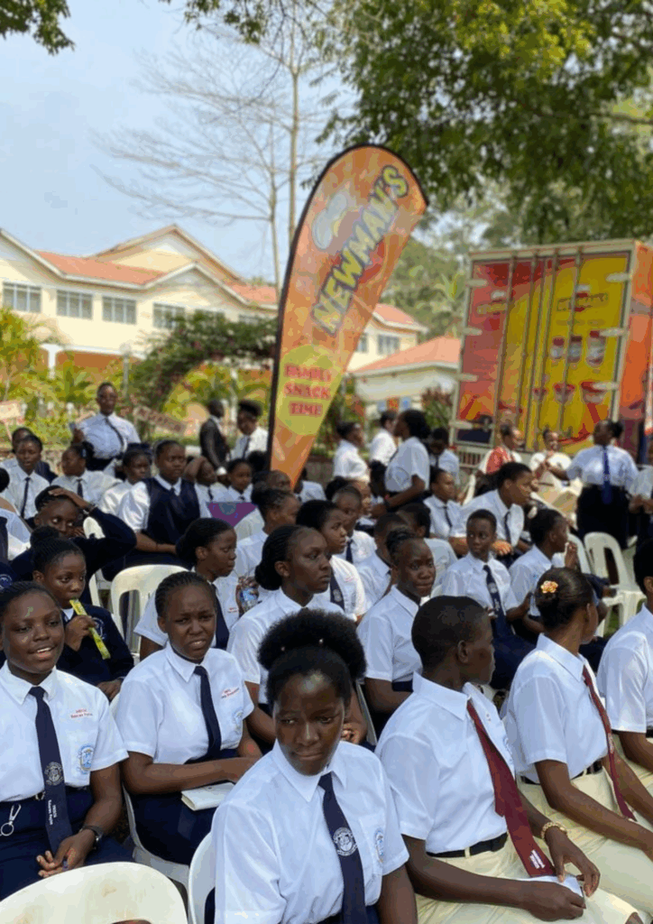 A large group of young women, dressed in school uniforms (white shirts and dark ties), are seated outdoors during an assembly or event. A large, orange and red Newman's banner advertising "FAMILY SNACK TIME" and a branded delivery truck are visible in the background.