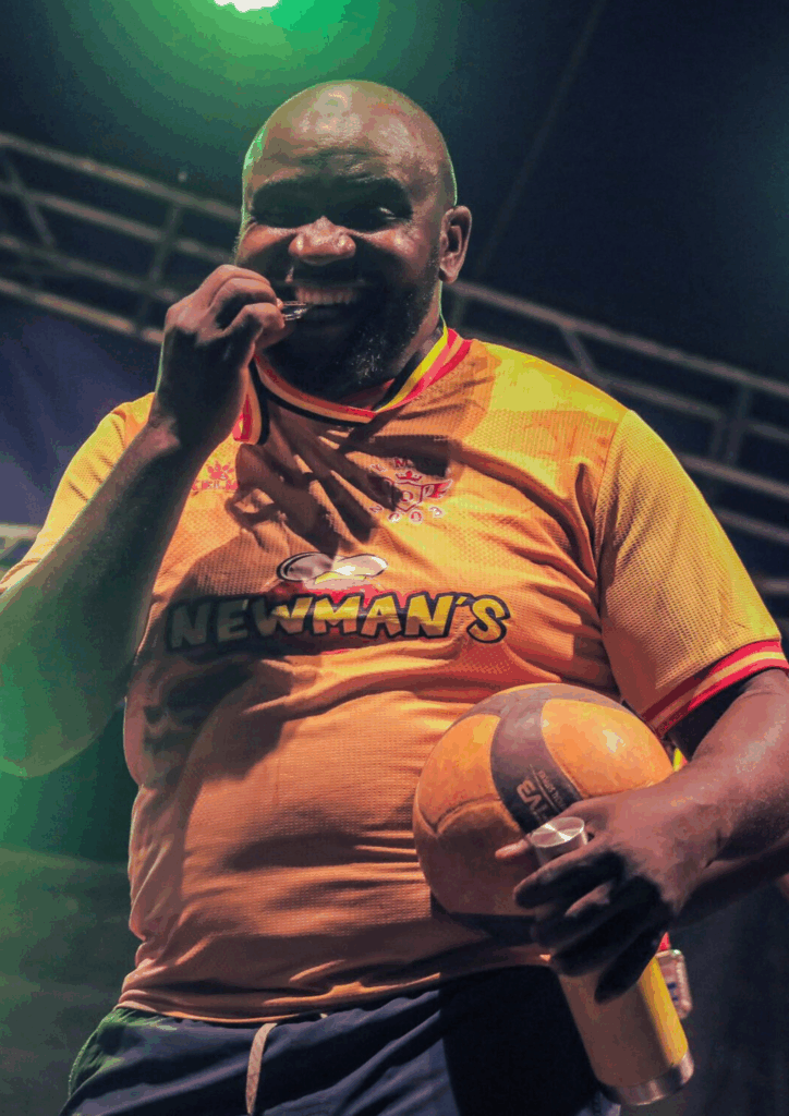 A man in an orange 'Newman's' branded jersey and a Ugandan flag patch, smiling while holding a volleyball at the KAMOSA Sports Gala.