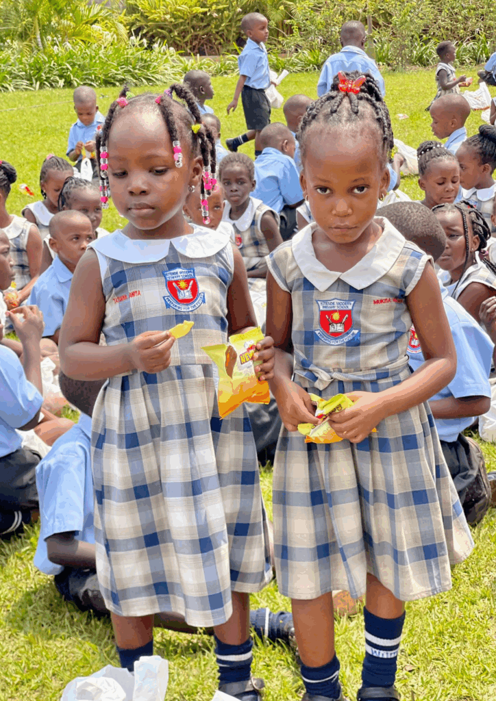 Two young female students in plaid school dresses stand holding bags of Newman's snacks, with many other students in uniform sitting behind them on the grass.