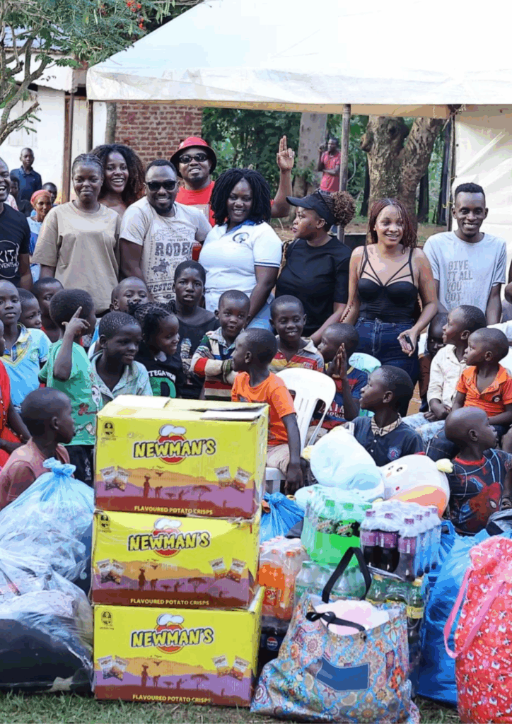 A large group photo of smiling adults (partners and Newman's staff) posing with dozens of children in front of a stack of Newman's crisps boxes and donated goods at Lighthouse Orphanage.