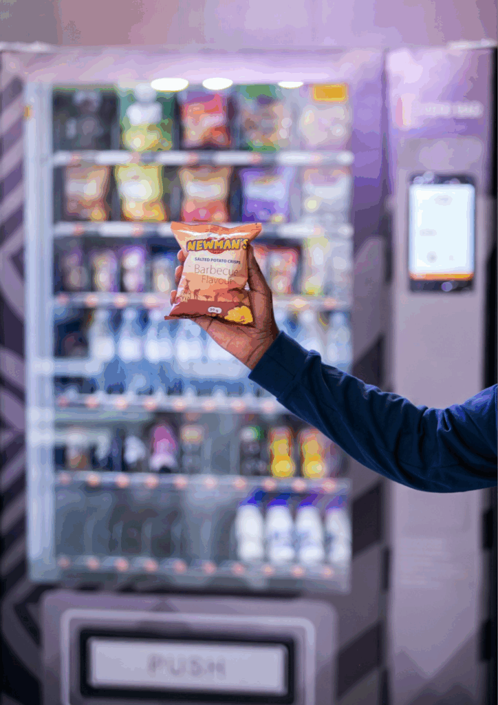 Hand holding a bag of Newman's Barbecue Potato Crisps in front of a vending machine, highlighting products on the Made in Uganda E-platform