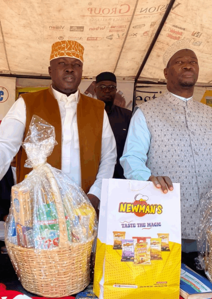 Three community leaders standing under a tent, presenting large gift baskets filled with Newman's products and other items to the Muslim community.