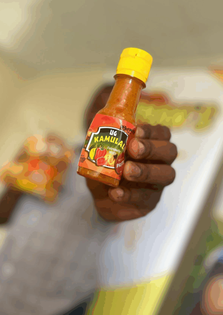 A hand, with the person out of focus in the background, holds up a small glass bottle of hot sauce. The bottle has a yellow cap and a label that reads "UG KAMULALI" at the top in black and yellow text, and "HOT PASTE" near the bottom. The label also features illustrations of chili peppers and tomatoes, and the words "EXTREMELY SPICY" are visible.
