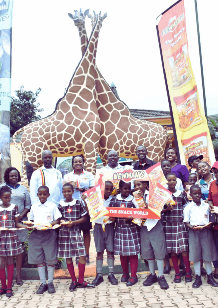 A group of adults (Newman's staff and UWEC officials) and smiling children in school uniform holding Newman's snack bags, posing in front of two large giraffe sculptures and a branded banner at the Uganda Wildlife Education Centre.