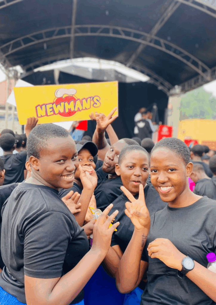 Three cheerful young women smiling and making peace signs in a large crowd in front of an outdoor stage with a Newman's banner.