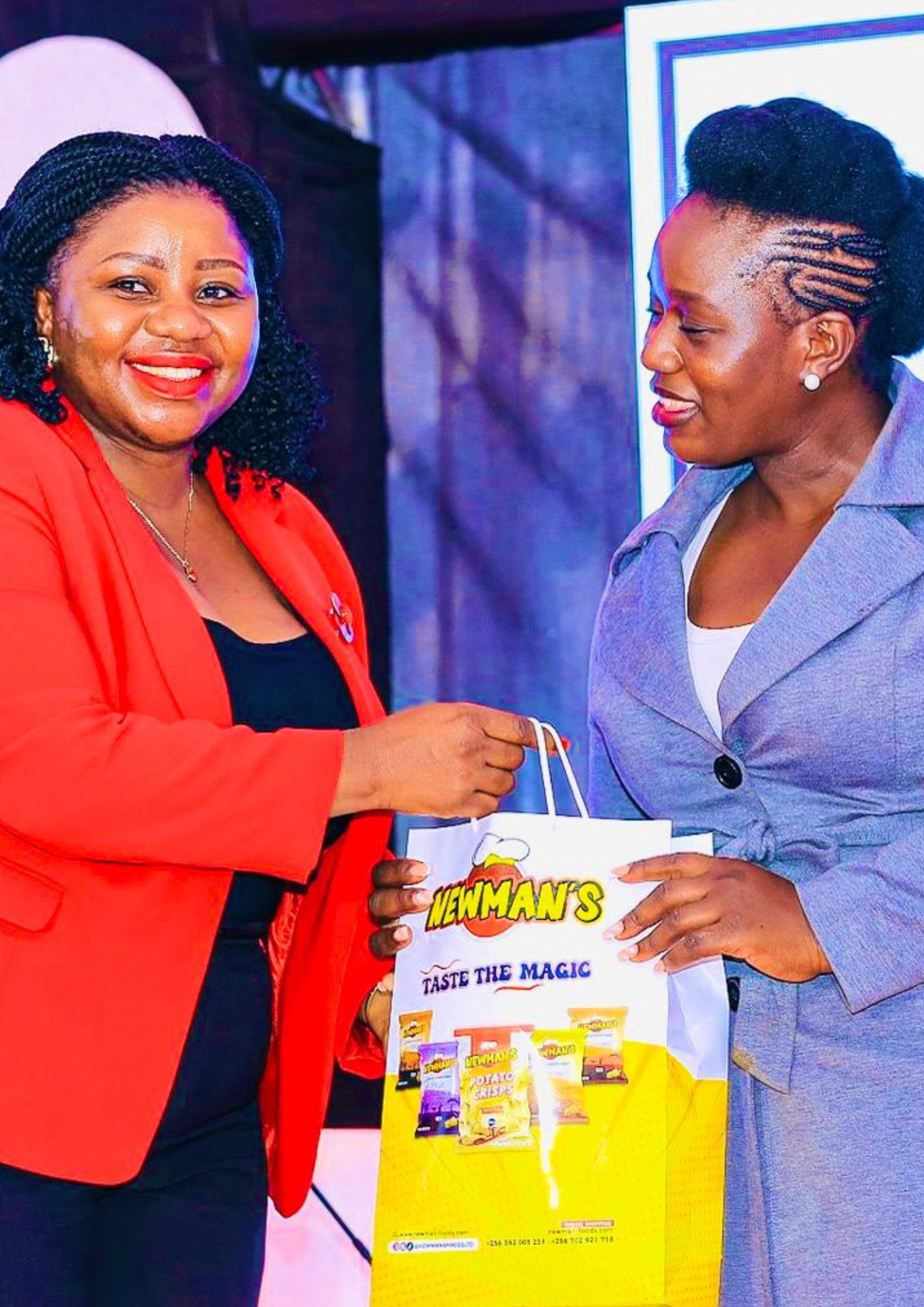 A woman in a red jacket smiles while handing a Newman's 'Bag of Goodness' to another woman in a grey coat on a stage, symbolizing professional recognition.