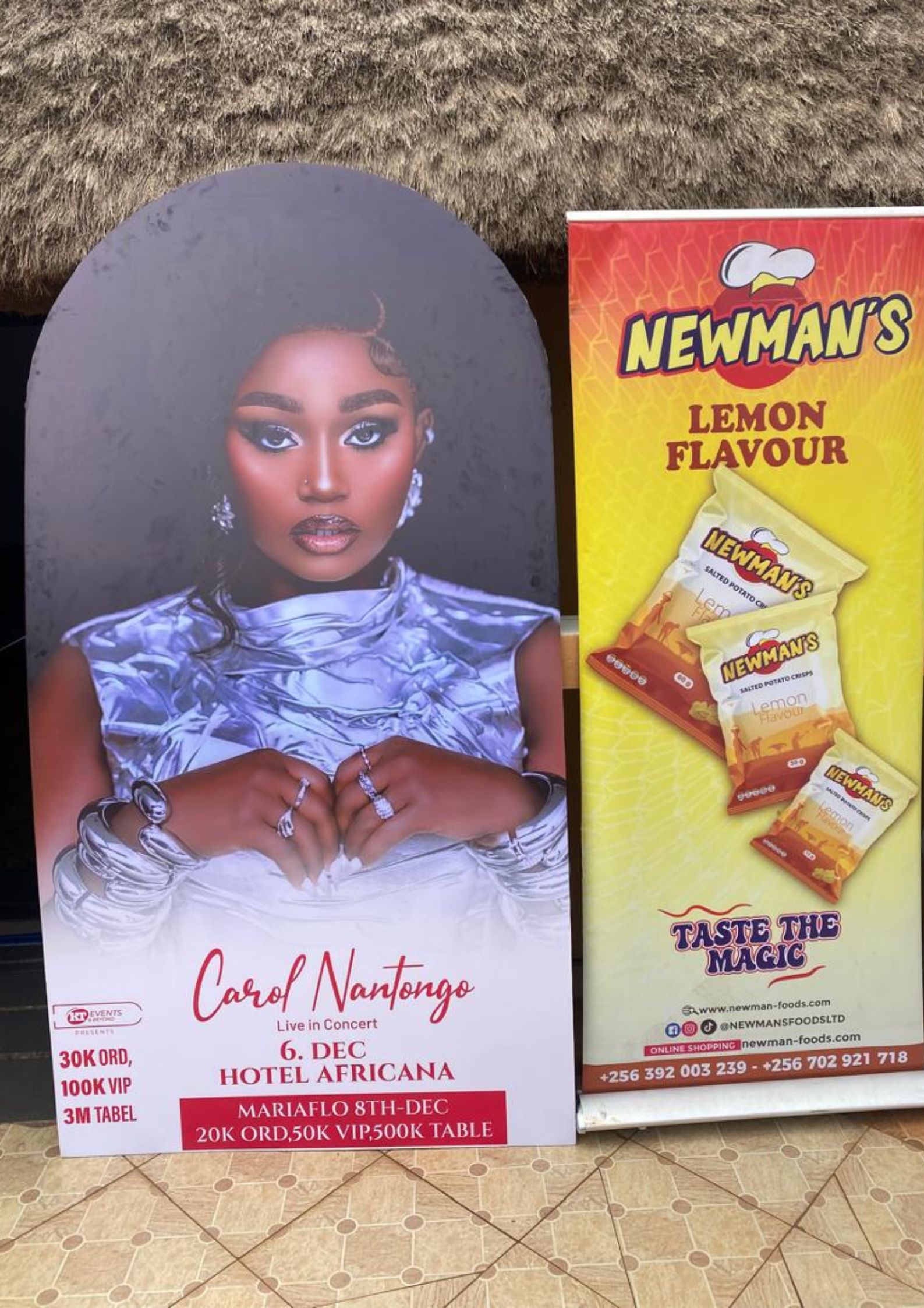 A full-size standee of Ugandan musician Carol Nantongo promoting her 'Live in Concert' show next to a tall, vertical banner for Newman's Lemon Flavour Potato Crisps.