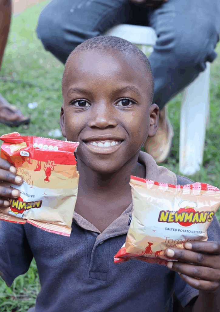 A close-up of a young Ugandan child smiling brightly while holding two small bags of Newman's Tomato Flavoured Salted Potato Crisps, with people seated in the background.