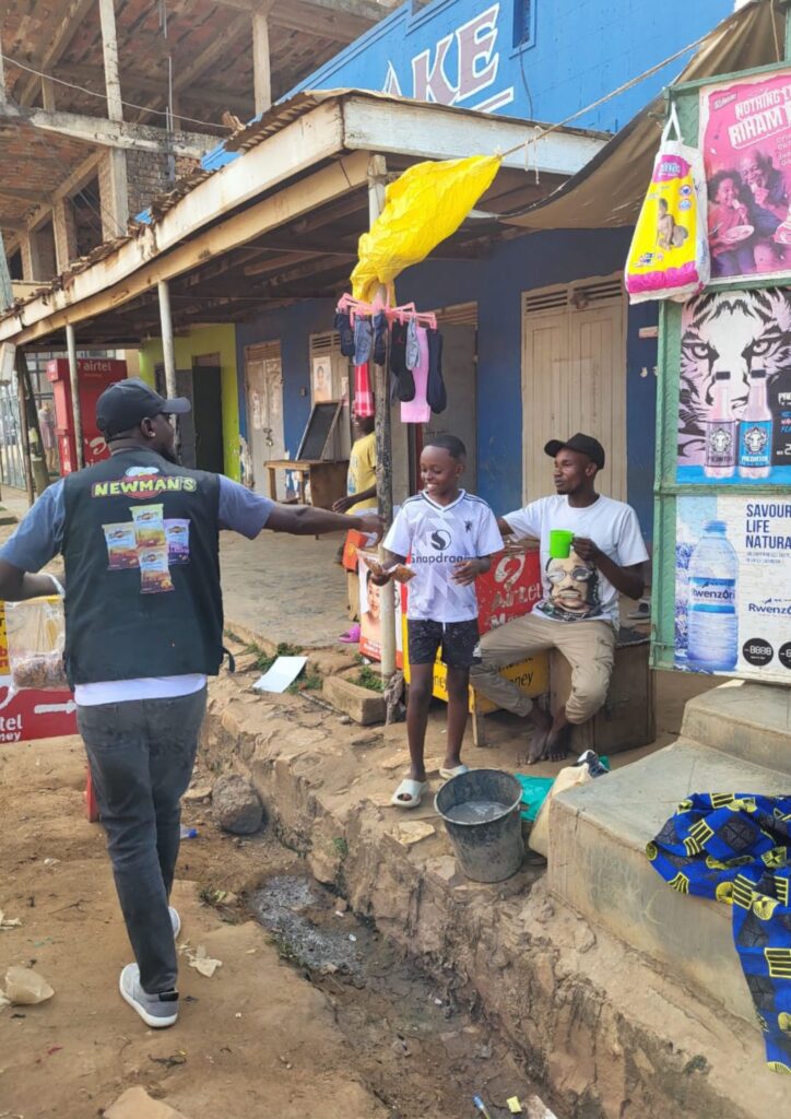 Newman's representative in branded vest handing snacks to a young boy and a seated man in a bustling roadside market in Iganga, Eastern Uganda, during a customer activation event.