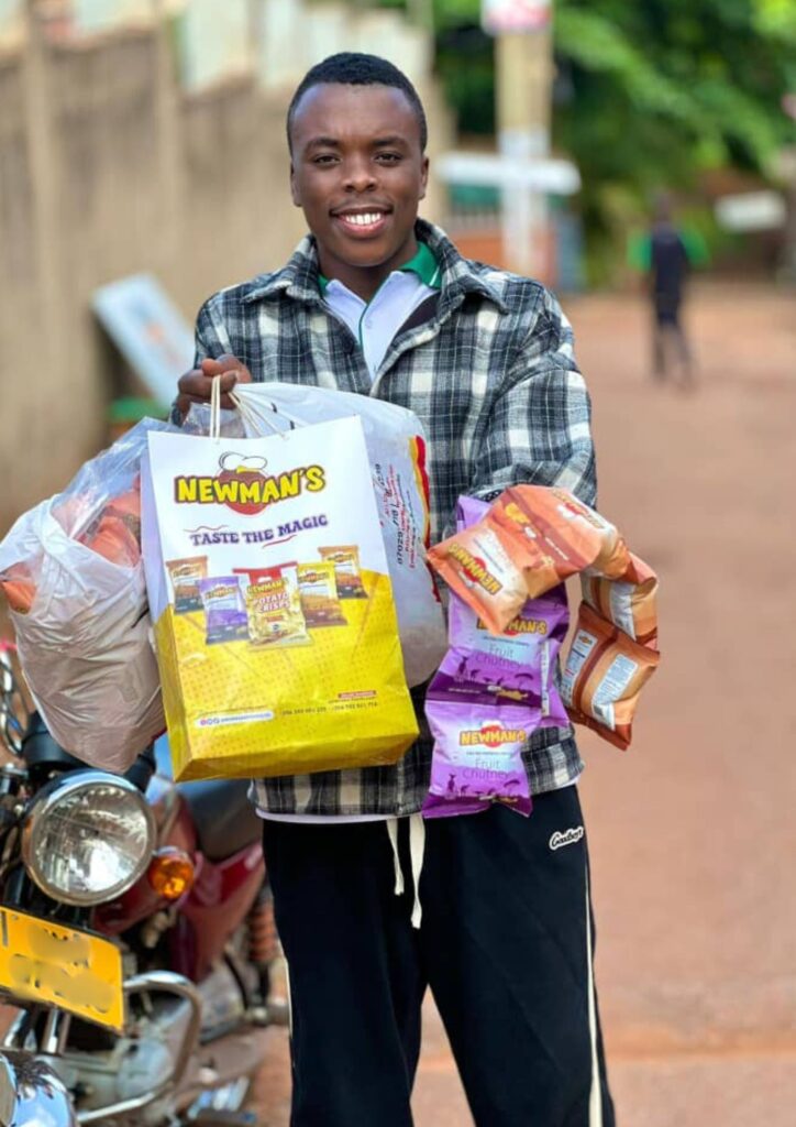 Influencer Ishmael smiling and standing next to a boda-boda, holding a large yellow Newman's "Taste The Magic" shopping bag and several clear bags filled with Newman's assorted snacks.
