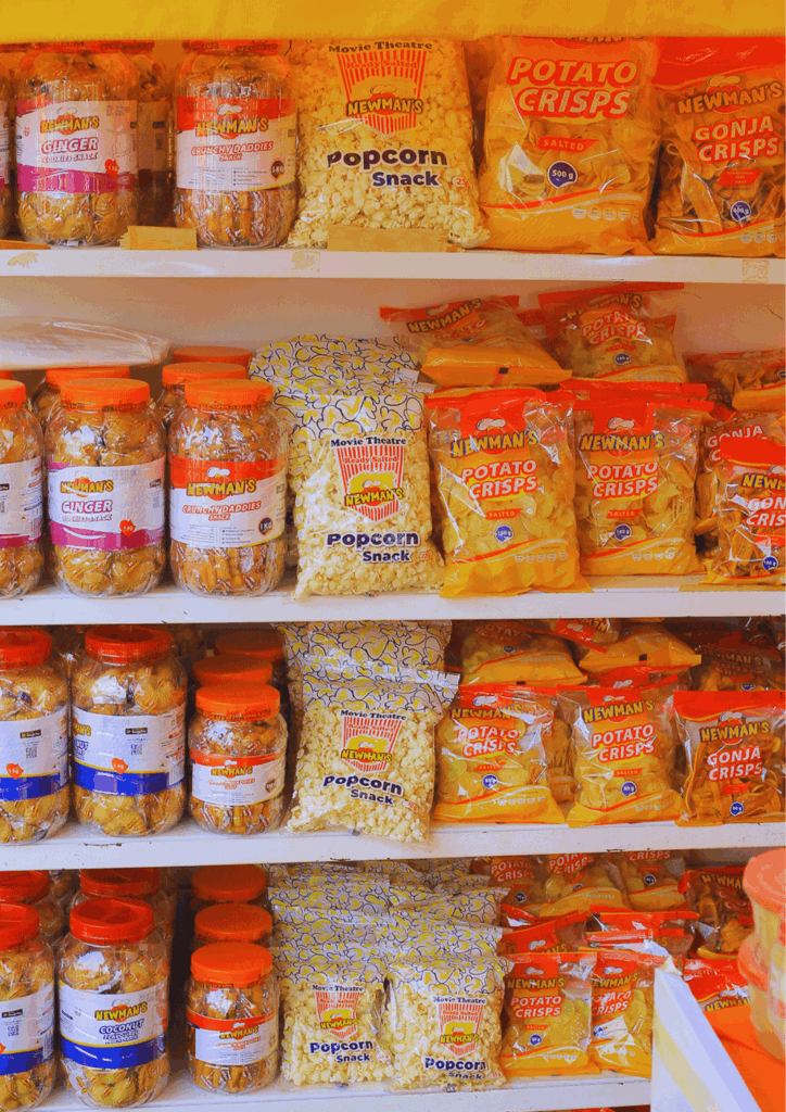 A shelf display fully stocked with jars of Newman's Ginger and Coconut cookies, large bags of Popcorn Snack, and smaller bags of Potato Crisps and Gonja Crisps, indicating high volume retail inventory.
