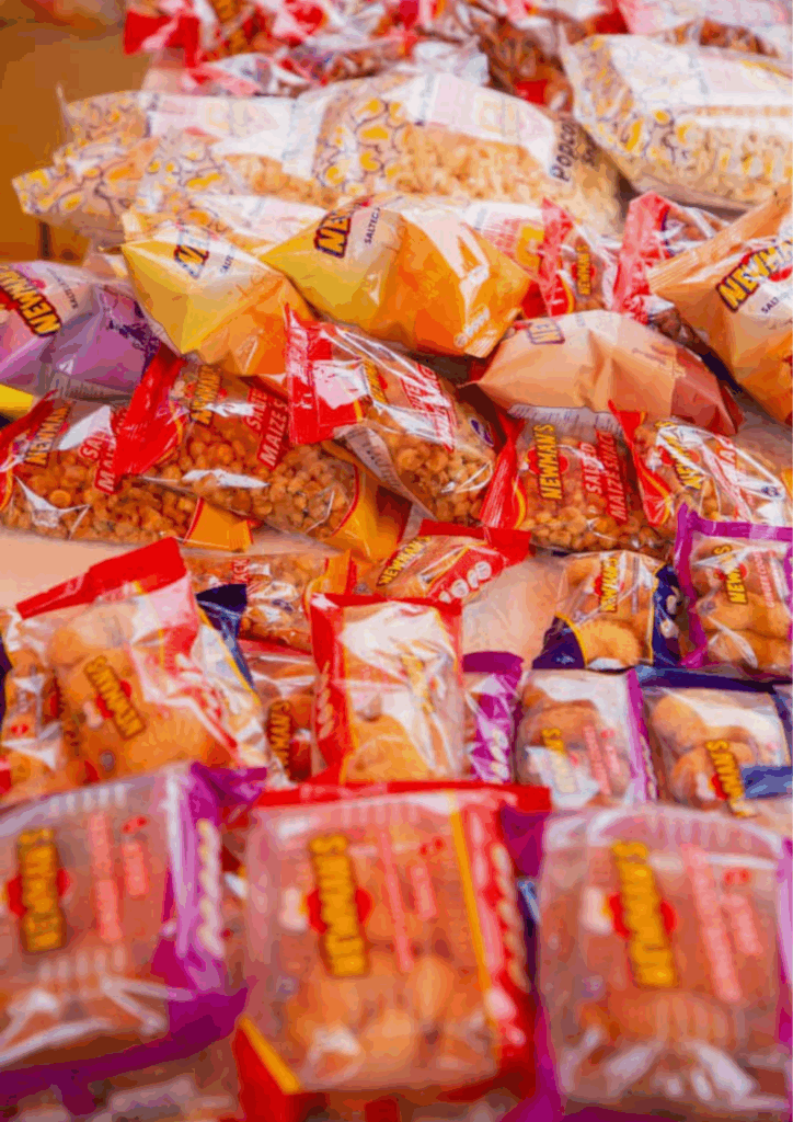 A close-up display of many individually packaged Newman's snacks, including crisps, maize puffs, and cookies, stacked for sale.