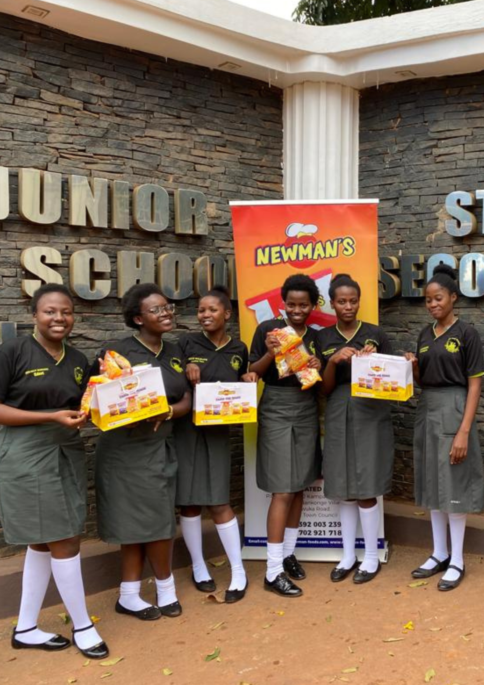 Five smiling female students in uniform outside St. Noa Girls Secondary School holding bags and boxes of Newman's snacks.