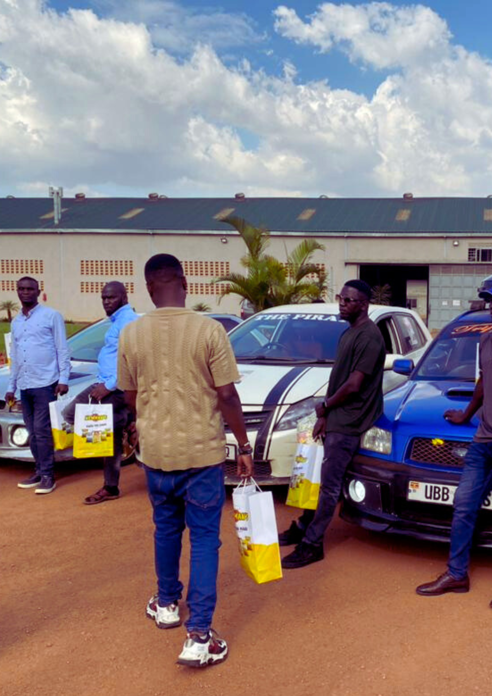 A group of drivers stand next to their performance cars, including a white and a blue Subaru, holding yellow Newman's gift bags at an event launch.