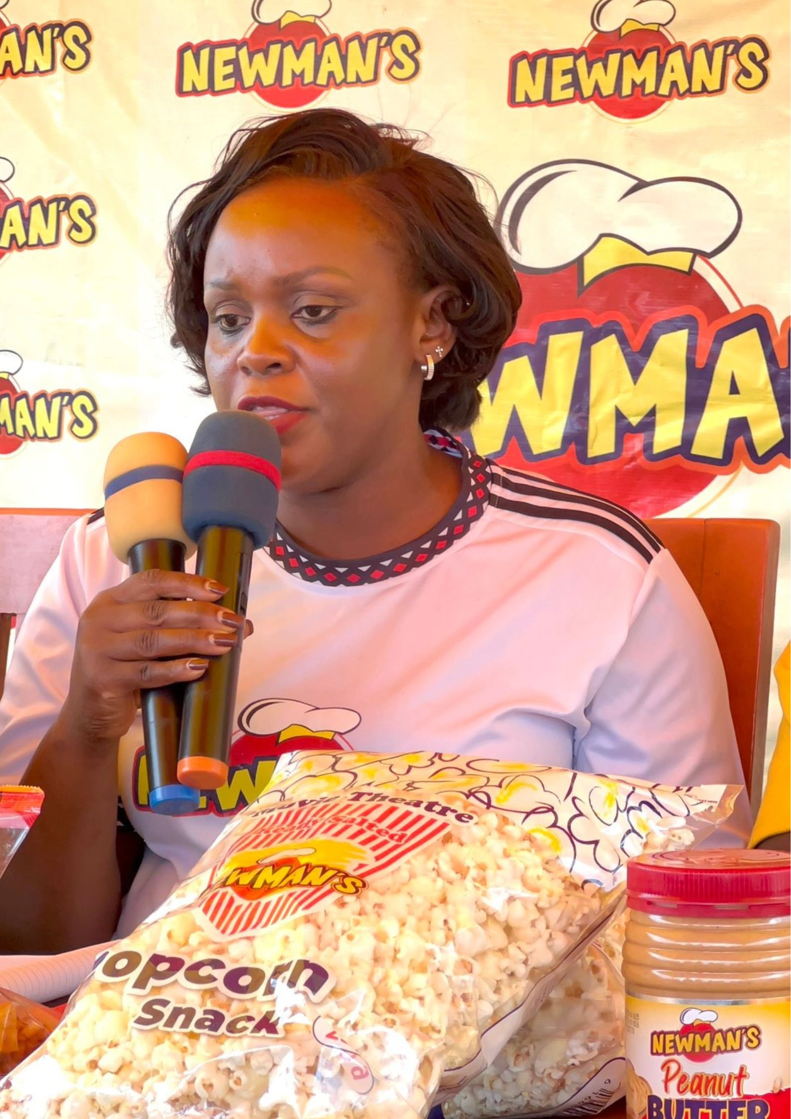 A close-up shot of Rosette Segujja speaking into two microphones, with the Newman's logo banner in the background and a large bag of Newman's popcorn on the table.